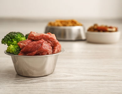 a metallic bowl in the foreground with raw meat and broccoli and two metallic bowls with food in the background